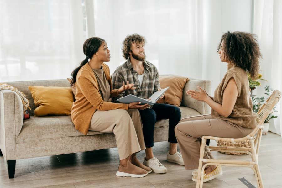 a young couple sitting on a couch with a booklet in hand speaking to a woman