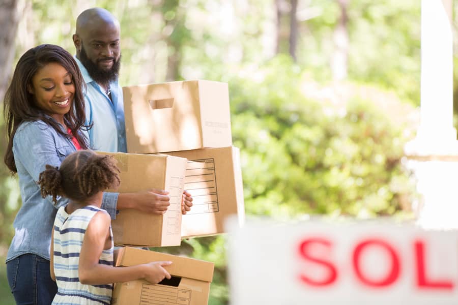 a couple and child carrying boxes in front of a sold sign