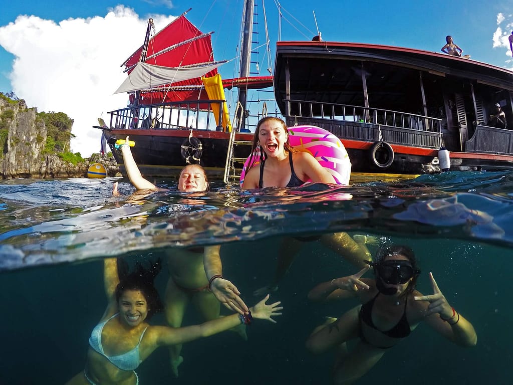 4 chicas buceando y nadando con la mitad de la foto mostrando escenas submarinas y la de arriba mostrando un barco pirata al fondo.