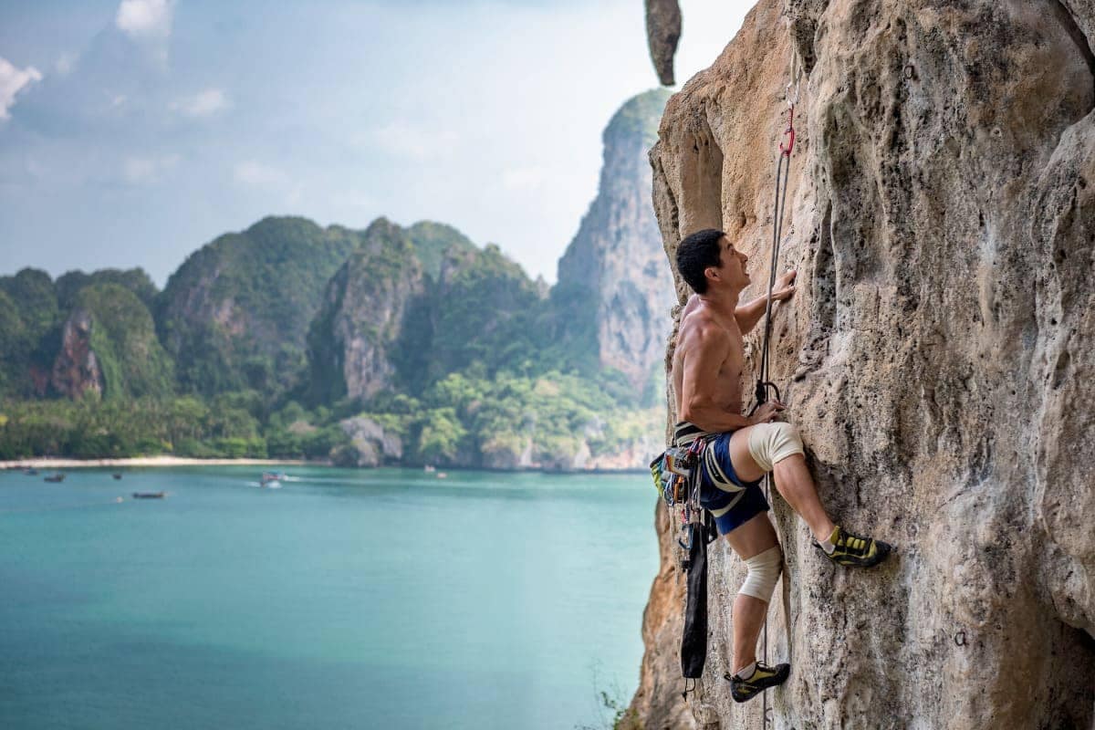 a man climbing a flat rockface with guide ropes, with Railay Beach and their limestone cliffs in the background to the left