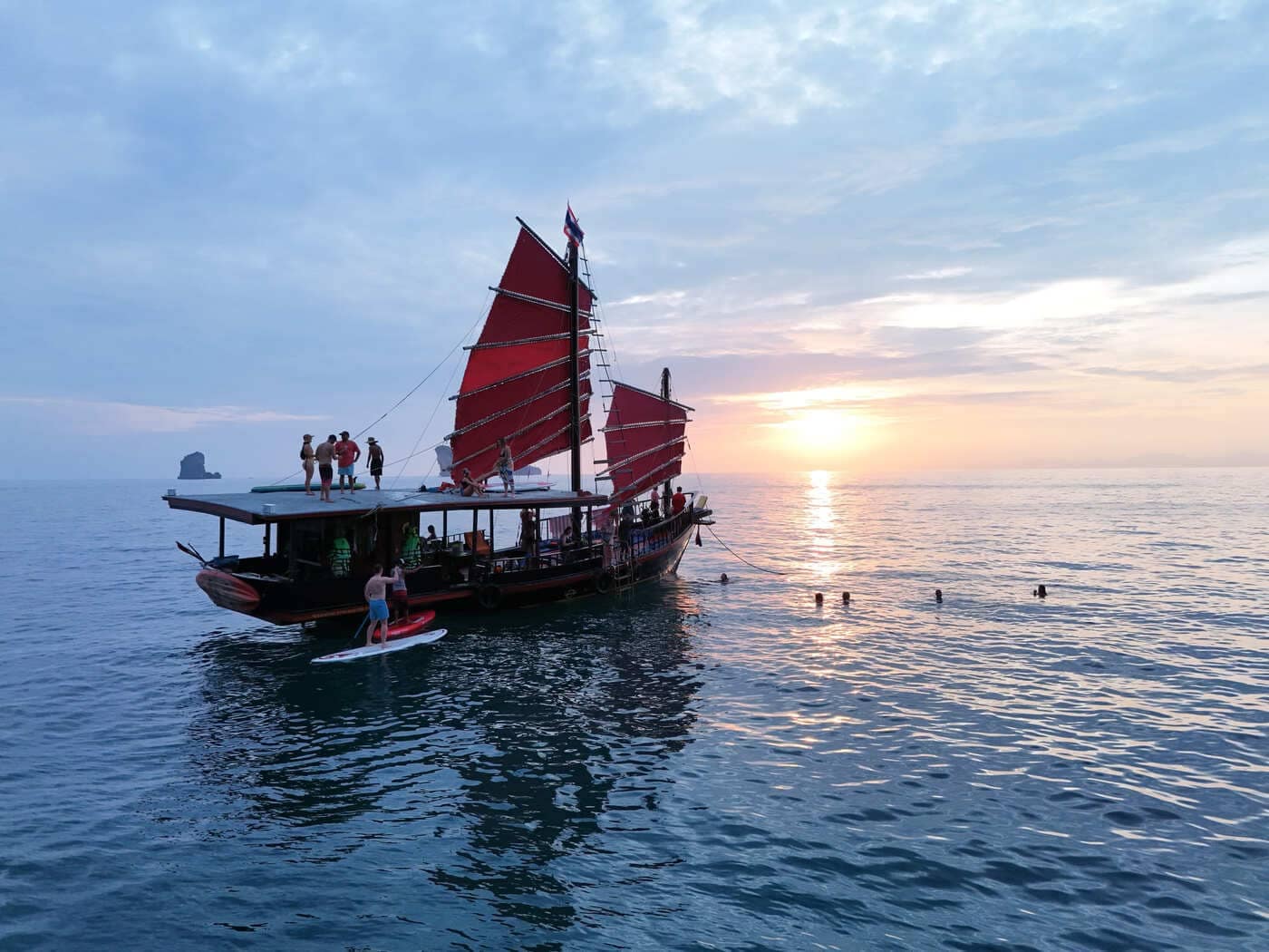 Barco tradicional siamés tipo «junk» al atardecer en Krabi, con gente practicando paddle surf y nadando a su lado.