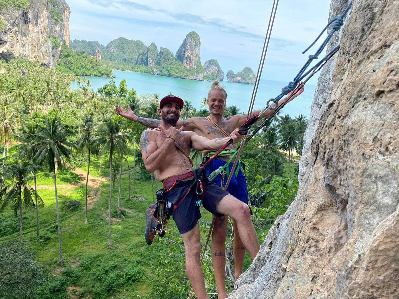 Dos escaladores posando en un acantilado de piedra caliza encima de la playa de Tonsai, en Krabi, con cuerdas de escalada, palmeras tropicales y el mar turquesa en el fondo.