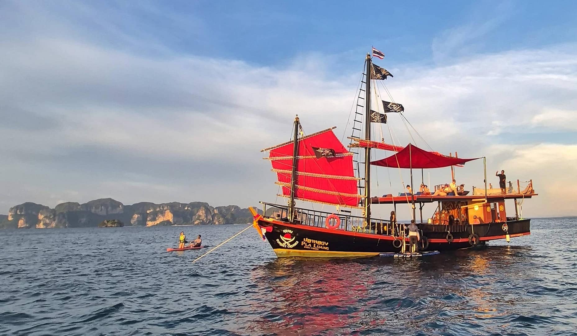 Krabi Sunset Cruises' traditional wooden Siamese junk boat with bright red sails, guests relaxing on the upper deck and kayaking beside the boat, with Krabi's limestone islands in the background.