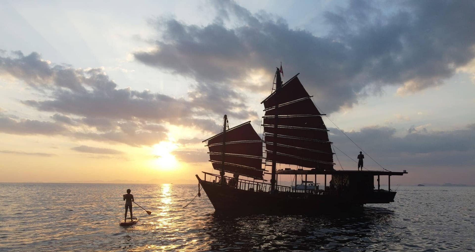 Crucero al atardecer en Krabi: un barco tradicional al atardecer con un practicante de paddle surf a su lado.