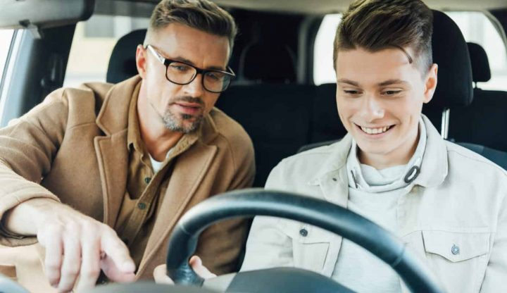 father pointing to something on the car dashboard while teaching smiling teen son driving car