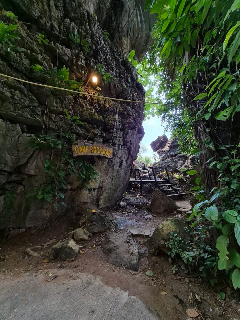 Entrada al Cave Rock Bar en la playa de Railay, con un cartel de madera, un camino de rocas, acantilados de piedra caliza y vegetación tropical.