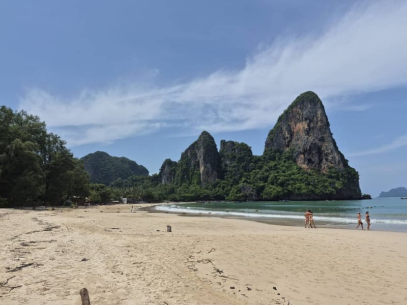 Playa de arena amplia en Railay West, en Krabi, Tailandia, con imponentes acantilados de piedra caliza, agua turquesa tranquila y visitantes que caminan a lo largo de la costa.