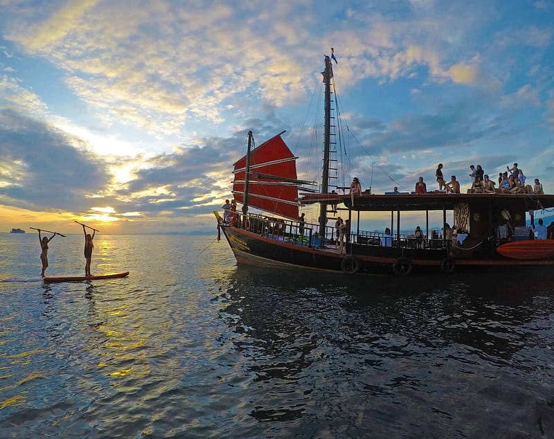 Krabi Sunset Cruises : une jonque au coucher du soleil, avec des passagers sur le toit et deux personnes sur des planches de paddle.