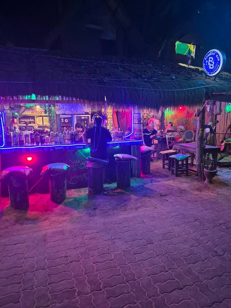 El Bamboo Bar en Railay Beach por la noche, con su techo de paja, iluminación colorida con neón, taburetes de bar de madera y clientes disfrutando de sus bebidas.