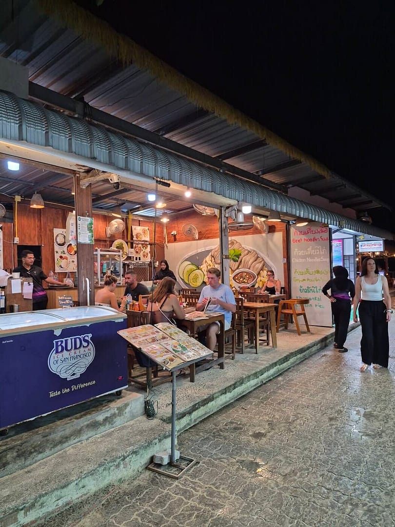 Turistas comiendo en un restaurante tailandés de comida rápida en Railay Walking Street por la noche, con mesas de madera, menús de comida y un área de asientos al aire libre.