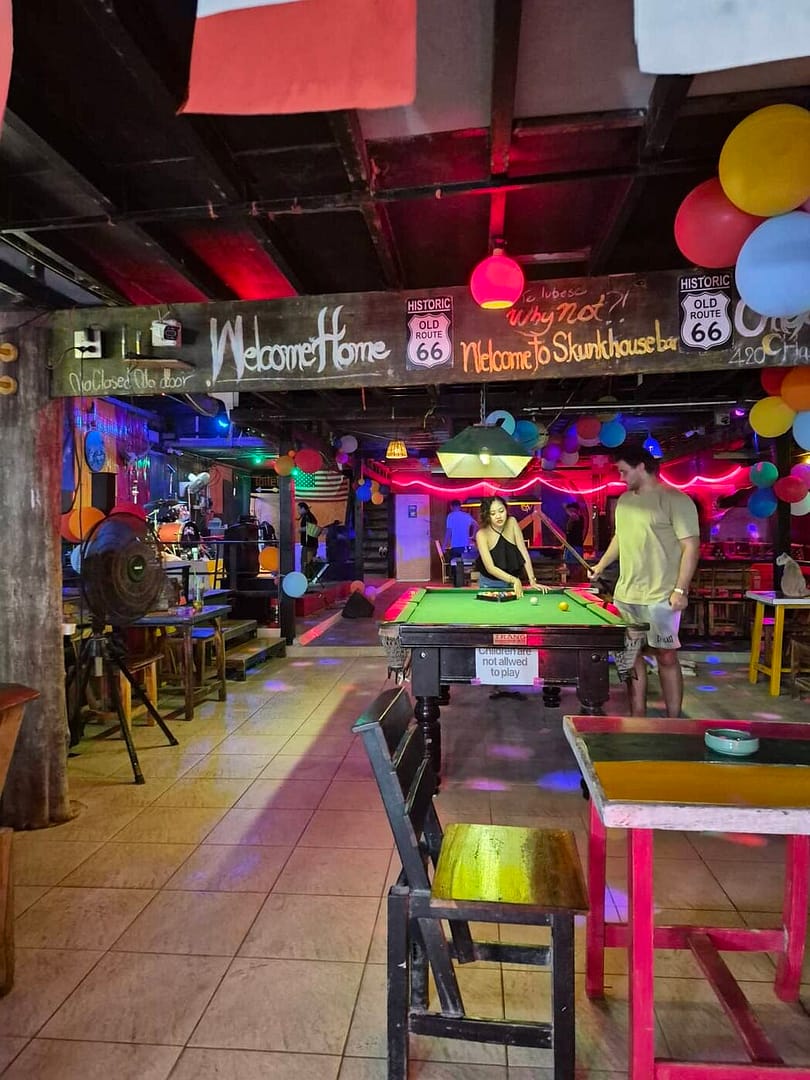 Dentro del Skunkhouse Bar en la playa de Railay, en Krabi, con visitantes jugando a billar, decoraciones coloridas y una iluminación neón brillante.