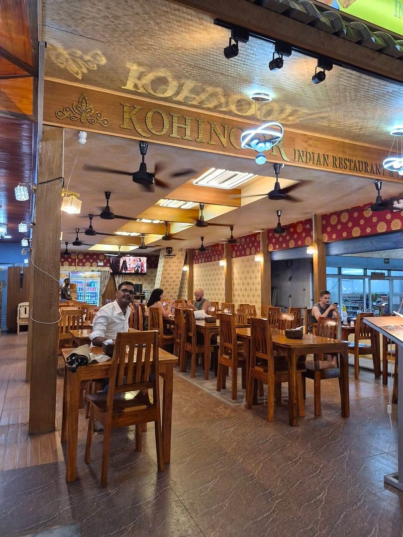 El interior del restaurante indio Kohinoor en la playa de Railay, en Krabi, con mesas de comedor de madera, ventiladores de techo y clientes disfrutando de la cena.