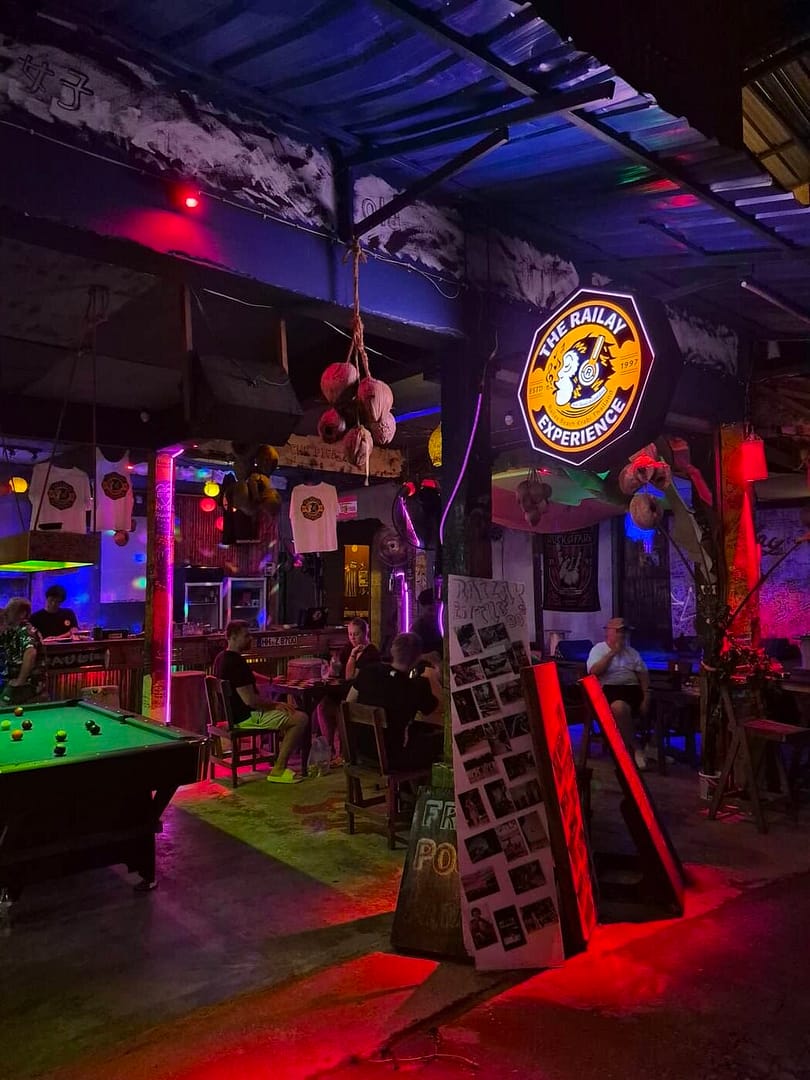 El interior del bar The Railay Experience por la noche, con una mesa de billar, iluminación colorida, asientos de madera y clientes relajados.