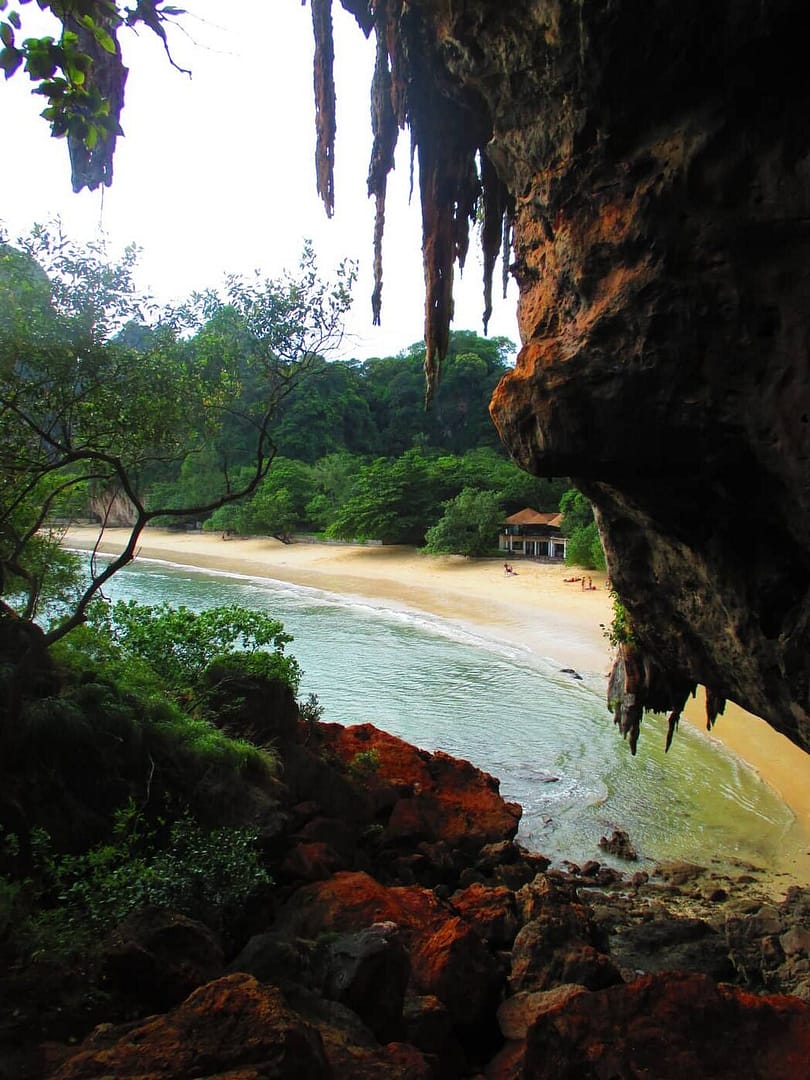 Playa de arena y agua turquesa vista desde el interior de una cueva de piedra caliza en la playa de Phra Nang, con formaciones rocosas colgantes y árboles tropicales.