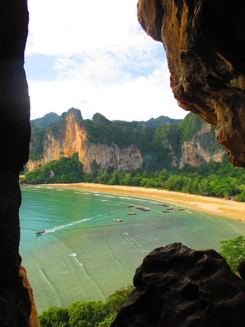 Vista de la playa de Railay West desde una cueva de piedra caliza que da a un mar de agua turquesa, barcos de pesca y acantilados imponentes en Krabi, Tailandia.