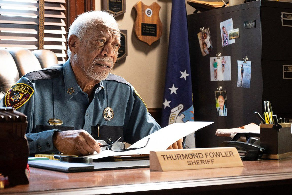 An old man (Morgan Freeman) in a police officer outfit seated behind a table holding a piece of paper.
