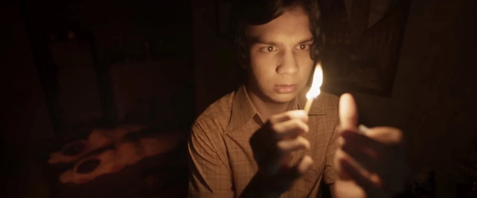 A young man holds a candle in a dark room with two bodies wrapped in blankets in the background.