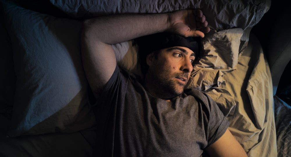 A man in a gray tshirt (Jason Schwartzman) and a black headband over his head peers over at a nightstand with a light shining overhead.