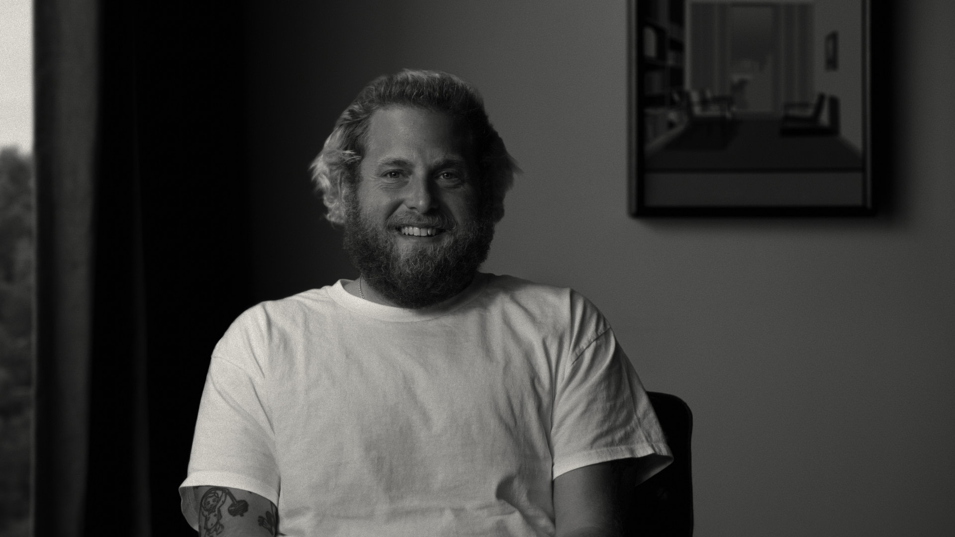 A black and white image of a bearded man (Jonah Hill) smiling and sitting in a chair against a blank wall with a portrait of an office hanging.