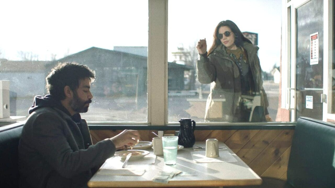 A man (Rahul Kohli) in a dark hoodie sits at a diner while a woman in sunglasses (Katie Parker) and a jacket taps at the window.