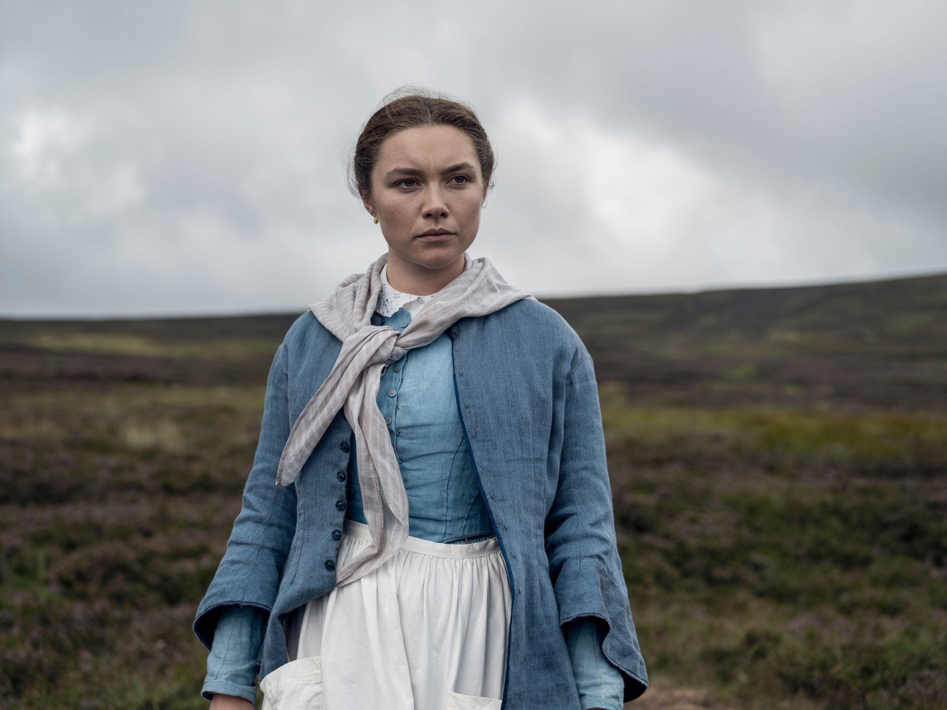 A nurse (Florence Pugh) in a blue and white dress with a white scarf standing in a low field of grace with a gray sky overhead.