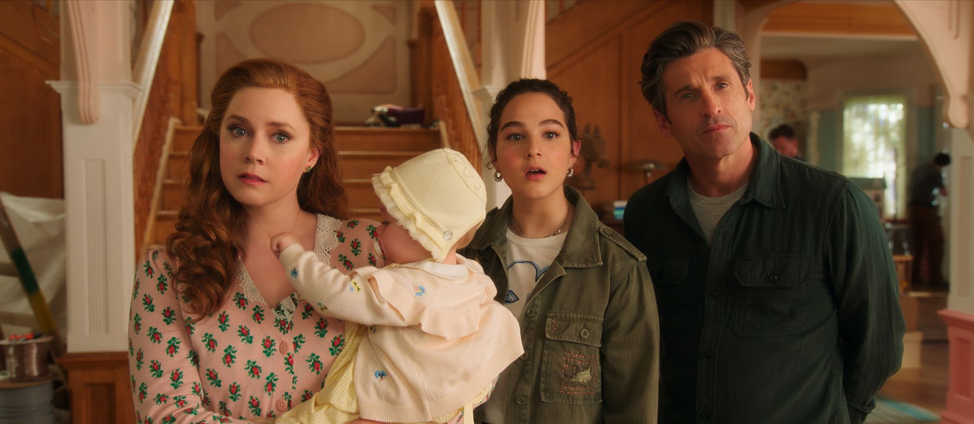 A red-haired woman in a pink floral shirt (Amy Adams) holds a baby beside a young girl in a green coat (Morgan Philip) and a man (Patrick Dempsey) while standing in front of a staircase and living room.