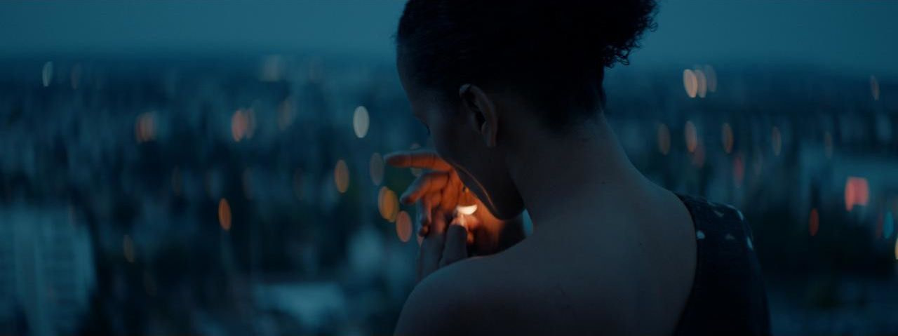 A woman with her back turned to the camera (Clarisse Albrecht) lights a cigarette in front of an out-of-focus cityscape at night.
