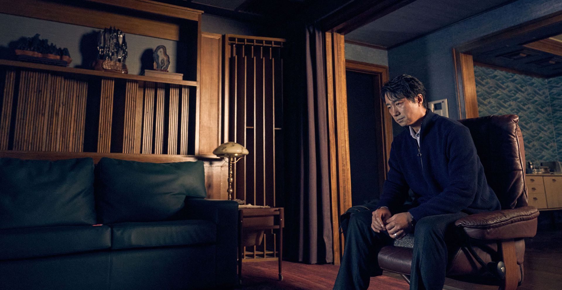 Detective Hae-jun (Park Hae-il), wearing a suit and looking exhausted, sits alone in a dark room lined with dark wood inlay in Decision To Leave