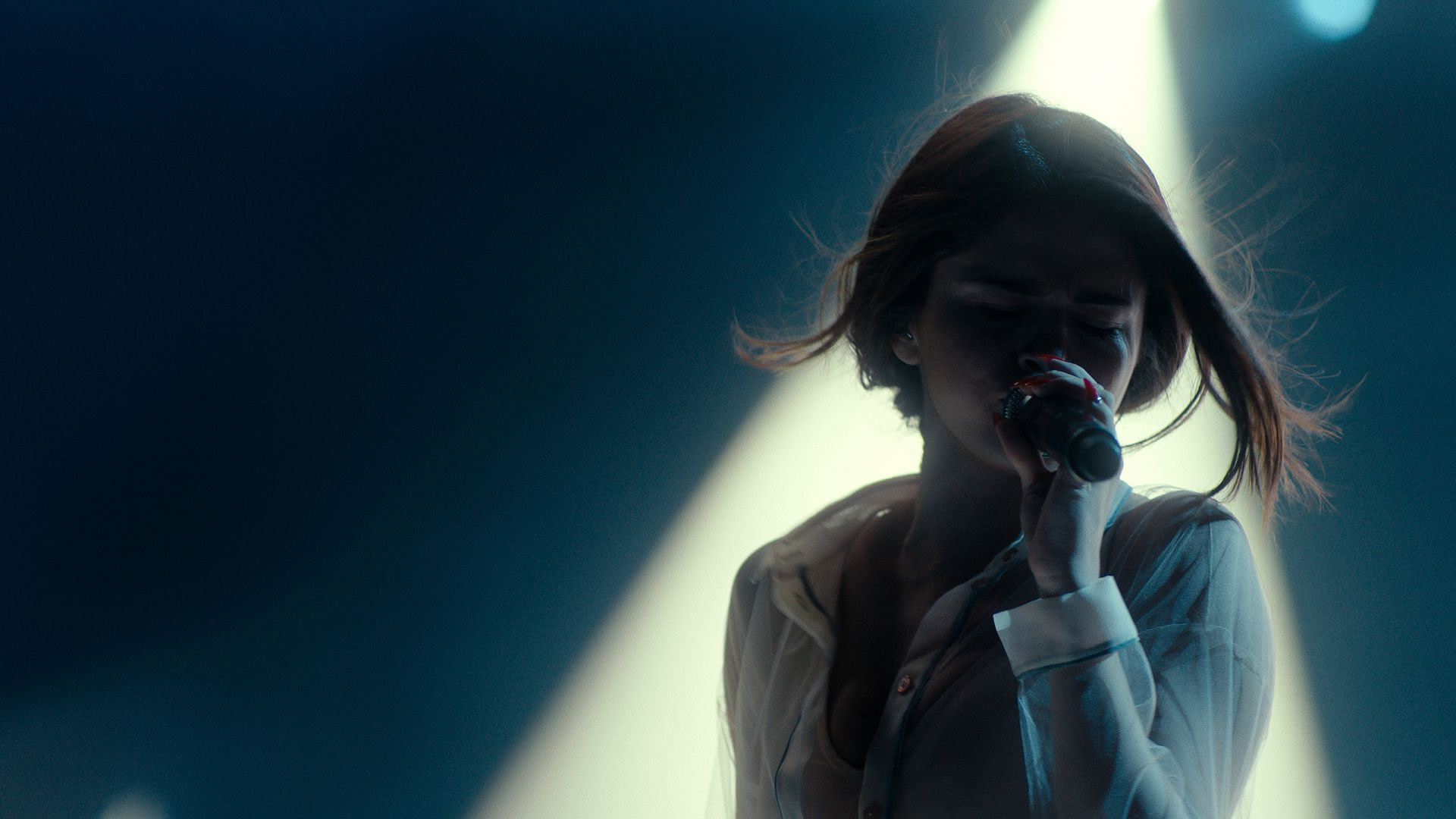 A woman (Selena Gomez) holding a microphone in one hand and singing while illuminated by a shaft of light in the background.