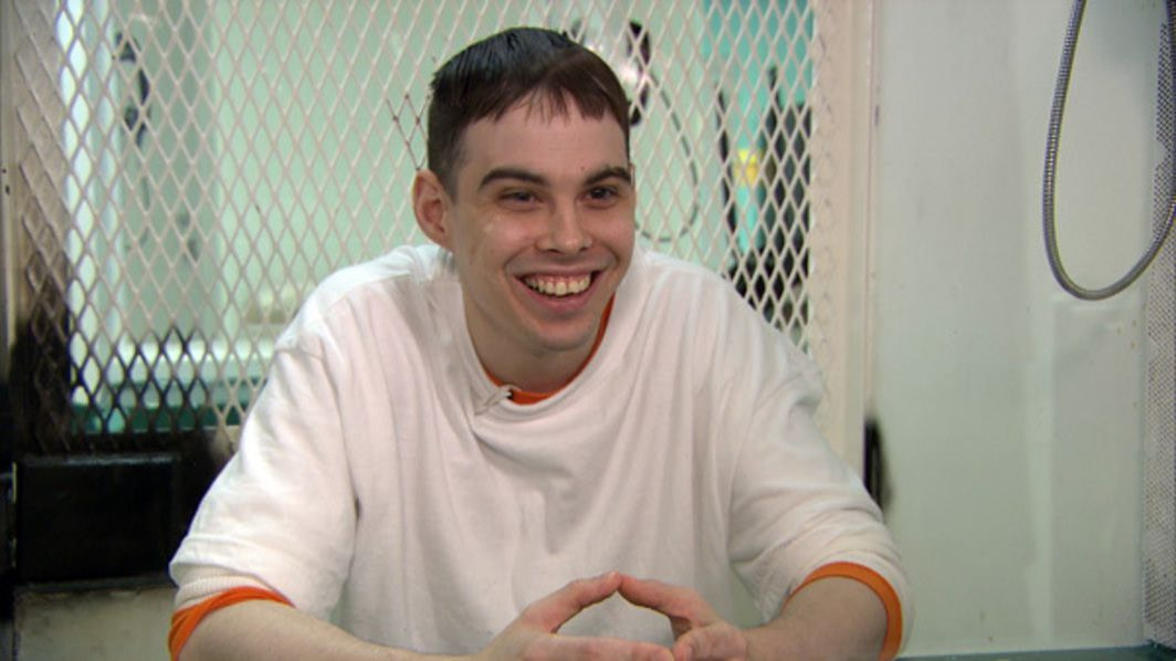 A smiling teenager with short-cropped brown hair in a white long-sleeved t-shirt over an orange t-shirt with its sleeves rolled up sits at a table with his fingers steepled in front of a window covered with white grating and black spray paint in the corner.