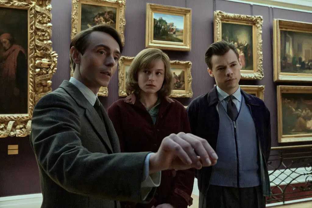A woman (Emma Corrin) in a burgundy coat stands between two men (L-R David Dawson, Harry Styles) in a museum as one of them gestures to something off-screen.