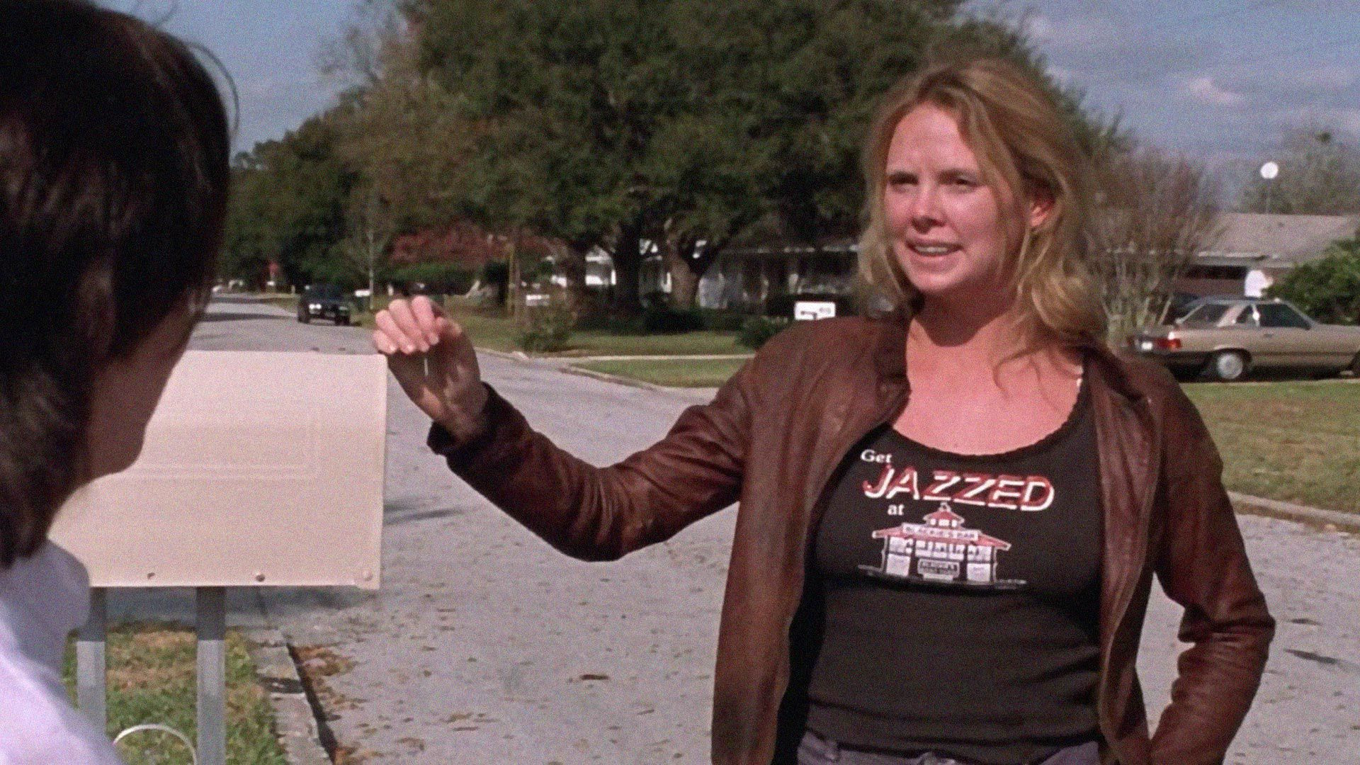 Charlize Theron in Monster, resting her hand on a mailbox while talking to someone. She wears a shirt that says “Get JAZZED at” with a picture of an establishment.