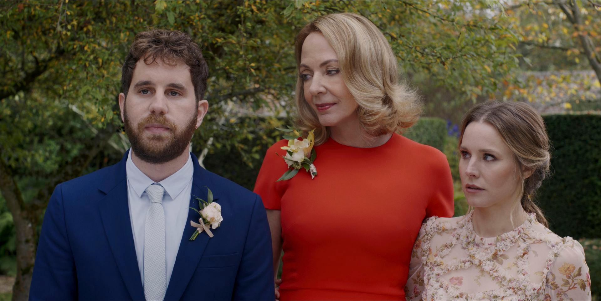 A man in a blue suit with a pink floral corsage (Ben Platt) stands next to a woman in a red dress with a yellow floral corsage (Allison Janney) and a woman in a pink floral dress (Kristen Bell) with a forest of trees and hedges in the background.