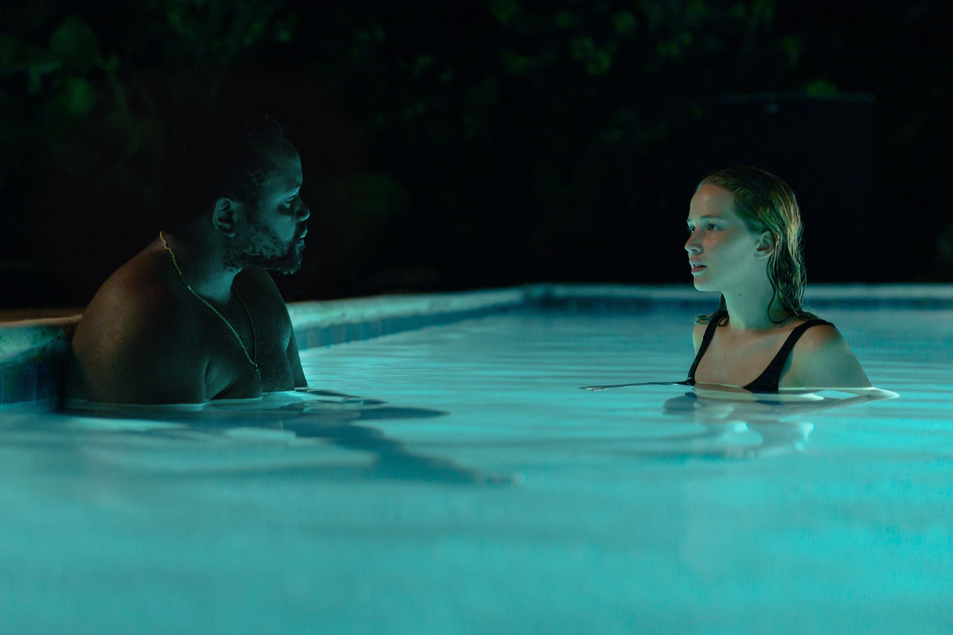 A man (Brian Tyree Henry) sitting in an illuminated pool across from a woman (Jennifer Lawrence) swimming at night-time.
