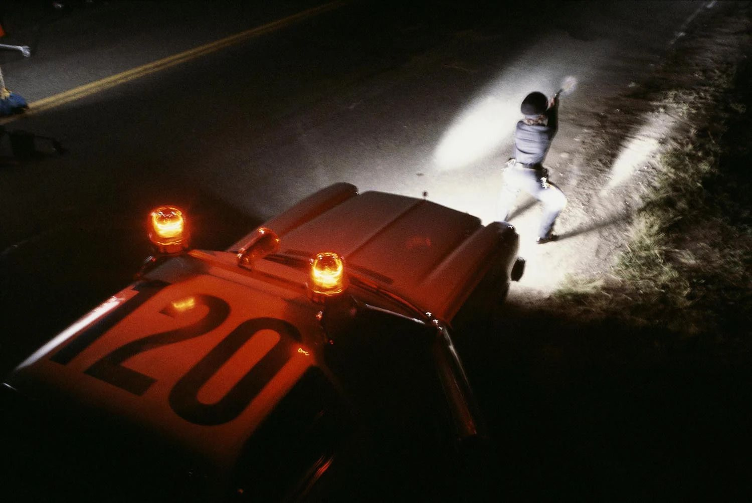 An overhead shot of a police officer wearing a cap squatting in front of a police cruiser at night, their back illuminated by the car’s headlights while aiming their pistol at something off-screen.