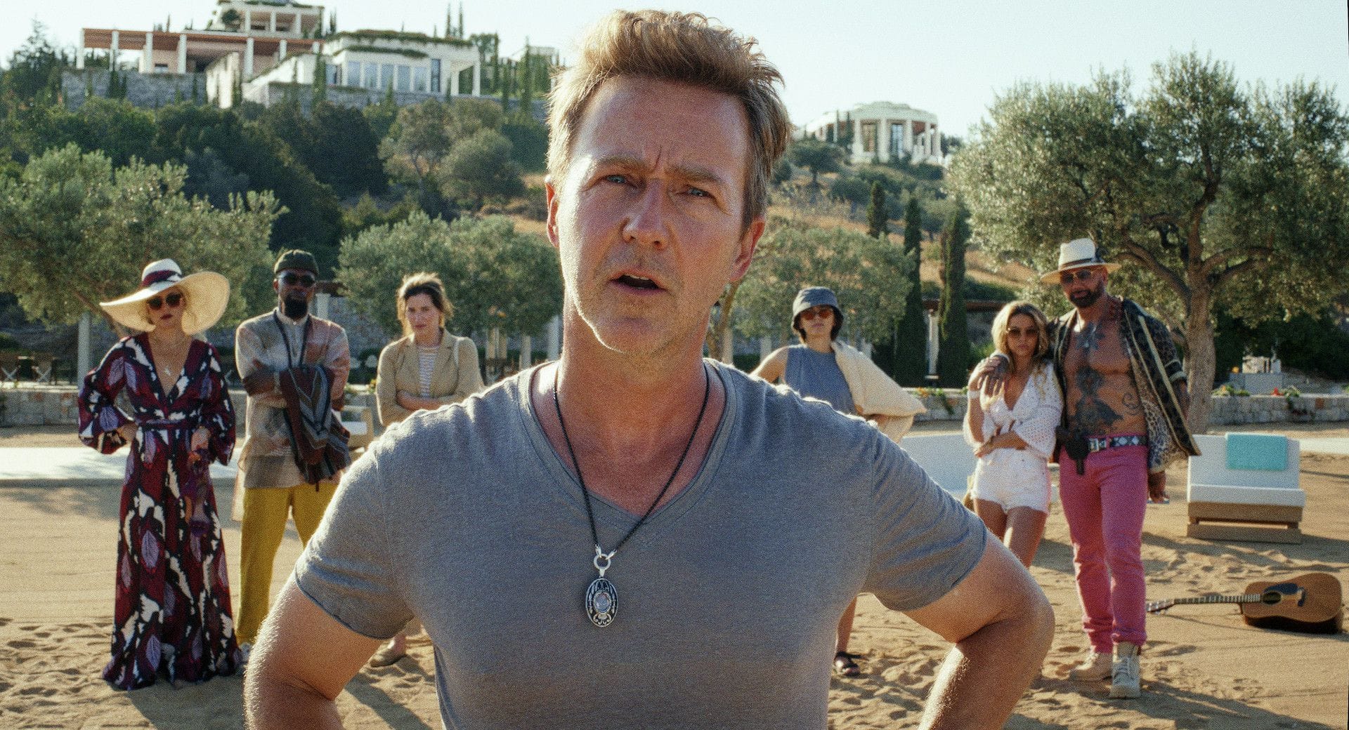 Edward Norton in the foreground, looking puzzled in a grey t-shirt on a Greek island beach, with an extravagantly dressed cast lined up behind him including Kate Hudson, Leslie Odom Jr, Kathryn Hahn, and Dave Bautista